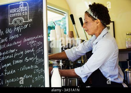 Une femme se tient devant un chariot de café mobile, prête à servir les clients. Cette scène d'affaires contemporaine met en valeur un café pop-up portable et l'esprit d'entrepreneur. Parfait pour les petites entreprises, la culture du café et les thèmes de l'hospitalité mobile. Banque D'Images