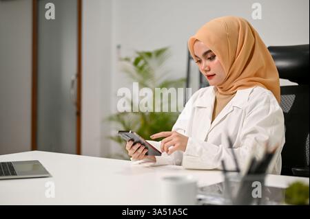 Une femme musulmane asiatique sérieuse portant un hijab utilise un smartphone à un bureau dans un bureau moderne, avec un ordinateur portable à proximité. Cette image convient aux thèmes des affaires, de la technologie et de la communication d'entreprise. Banque D'Images