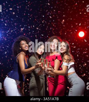 Groupe d'amies féminines diverses tenant des flûtes à champagne dansant dans une chambre sombre avec des lumières rouges et bleues et des confettis tombant pendant la Nouvelle Yearâs Eve p... Banque D'Images