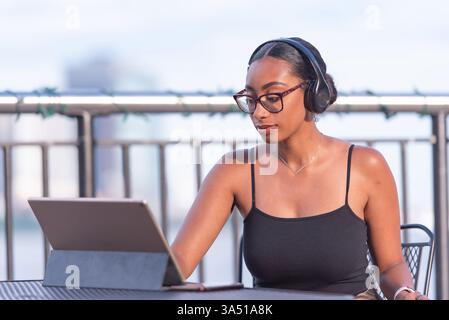Femme afro-américaine portant des lunettes et des écouteurs travaillant à une table dans un restaurant en plein air pendant la journée. Cette scène d'espace de travail décontracté convient aux campagnes de travail à distance, de freelance ou de culture de café. La lumière naturelle et le cadre extérieur ajoutent une ambiance détendue et productive. Banque D'Images
