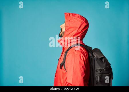 Vue latérale d'un touriste barbu dans une veste rouge avec un sac à dos. Sur fond bleu dans un studio lumineux, l'image transmet voyage et exploration. Idéal pour le style de vie, le voyage et le contenu d'aventure. Banque D'Images