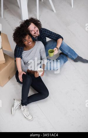 Joyeux couple multiethnique se relaxant dans une nouvelle maison, assis sur le sol avec du café comme des boîtes en carton les entourent. Image chaleureuse et authentique adaptée au déménagement, à la vie à la maison et aux campagnes immobilières. Transmet la convivialité, les nouveaux départs et les moments confortables de la vie quotidienne. Banque D'Images