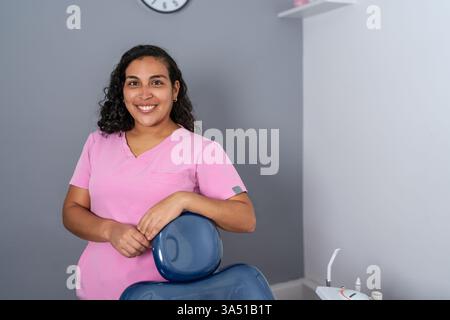 Dentiste hispanique souriante avec les cheveux bouclés appuyés sur une chaise dentaire dans une clinique moderne. Idéal pour les soins dentaires, la santé bucco-dentaire et les visuels professionnels de la santé. Banque D'Images