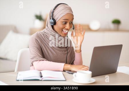 Happy Black Woman dans un hijab participe à une vidéoconférence sur son ordinateur portable, discutant avec des collègues et faisant signe devant la caméra. Cette scène de bureau à domicile met en avant le travail à distance, la collaboration en ligne et la communication professionnelle. Idéal pour les affaires, la technologie et les thèmes de diversité. Banque D'Images