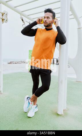 Homme hispanique portant des écouteurs se tient dans une salle de sport en plein air près de la plage, regardant loin avec les pieds croisés. Cette image de style de vie énergique convient au fitness, aux entraînements en plein air et aux loisirs en bord de mer. Banque D'Images