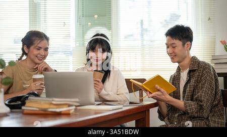 Groupe d'amis universitaires collaborant à un projet, à l'aide d'un ordinateur portable et d'un livre de référence. Ils s'assoient ensemble sur le campus à la recherche d'informations et de brainstorming d'idées. Parfait pour l'éducation, le travail d'équipe, la vie étudiante et les campagnes d'étude. Banque D'Images