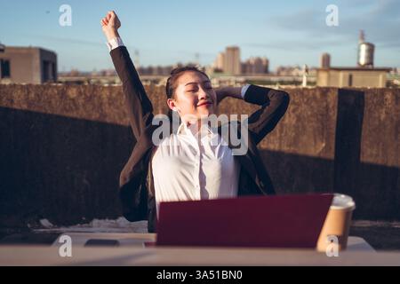 Femme asiatique assise à une table sur le toit avec un ordinateur portable, étirant ses mains. Cette scène moderne de travail à distance convient aux histoires de travail flexible et de style de vie urbain. Parfait pour la technologie, l'entrepreneuriat ou les visuels de bien-être au travail. Banque D'Images