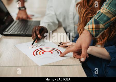 Une équipe diversifiée de collègues collabore sur un projet autour d'un bureau, avec un ordinateur portable et des graphiques. Les mains pointent vers les données, soulignant le travail d'équipe, la stratégie et un environnement de bureau moderne. Banque D'Images