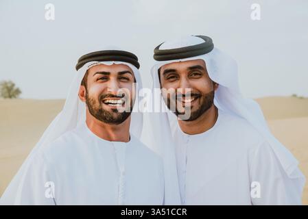 Des hommes arabes souriants portant du kandura se tiennent ensemble dans un décor désertique. Idéal pour la culture du moyen-Orient, le réseautage d'affaires et les visuels de style de vie régional aux Émirats arabes Unis. Banque D'Images