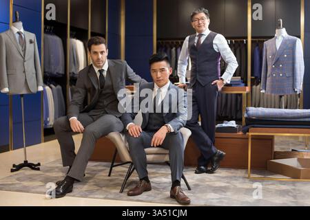 Un créateur de mode chinois se tient la main dans sa poche aux côtés d'un groupe diversifié d'hommes d'affaires chinois et hispaniques assis ensemble dans un studio de design. L'image exprime le leadership, l'expertise en design de mode et la collaboration dans un environnement moderne de vente au détail ou de studio. Idéal pour le contenu sur l'industrie de la mode, les vêtements de créateur et le travail d'équipe professionnel. Banque D'Images