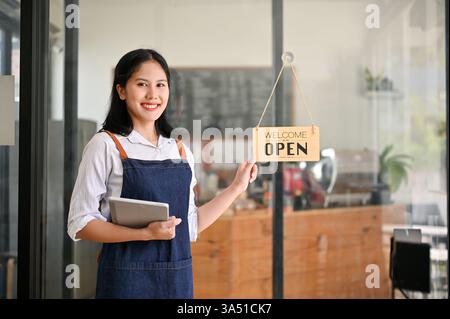 Sympathique jeune femme asiatique se tient à l'entrée du café avec une tablette numérique dans sa main. Un portrait accueillant de l'hospitalité adapté pour café, café, ou petite entreprise de marque. Idéal pour le marketing de restaurant, le service en front-of-House et les campagnes d'expérience client. Banque D'Images
