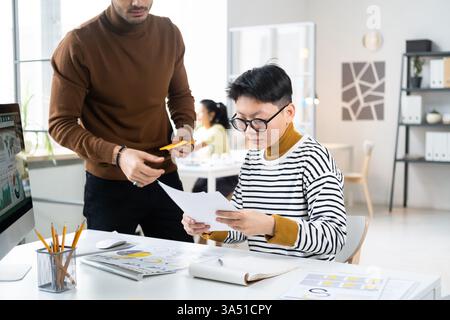 Homme d'affaires du moyen-Orient debout et parlant à une collègue asiatique examinant des documents tout en étant assis à table avec l'ordinateur près de collègues ... Banque D'Images
