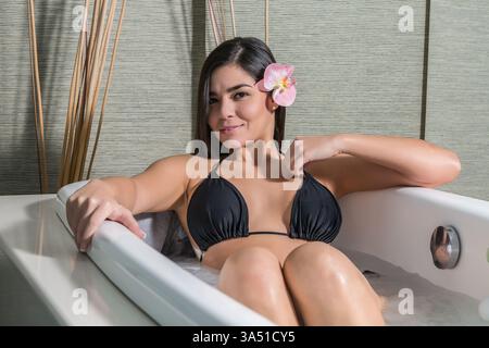 Une femme hispanique souriante en bikini se détend dans une baignoire spa. Le cadre lumineux et apaisant du spa met l'accent sur le bien-être et les soins personnels. Idéal pour les campagnes beauté, bien-être et lifestyle. Banque D'Images