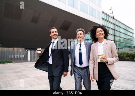 Groupe diversifié marchant près d'un bâtiment de la ville, mettant en vedette une femme hispanique et des collègues masculins caucasiens. La scène communique la collaboration et le travail d’équipe professionnel dans un environnement urbain. Idéal pour les entreprises, le marketing de diversité et les communications d'entreprise. Banque D'Images