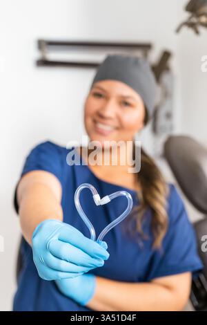 Sourire dentiste mixte femme portant des gants forme une forme de coeur avec des instruments dentaires dans une clinique. Cette image professionnelle fonctionne bien pour la santé, la dentisterie et les campagnes de soins aux patients. Banque D'Images
