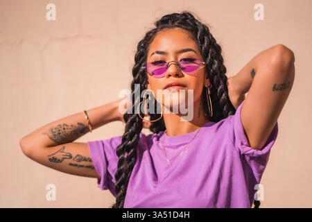 Urban session montre une jeune femme à la peau foncée avec de longues tresses et des lunettes violettes, debout avec un regard sérieux sur un fond Uni. Le portrait respire l'attitude et le style confiant dans un contexte de style de vie urbain. Banque D'Images
