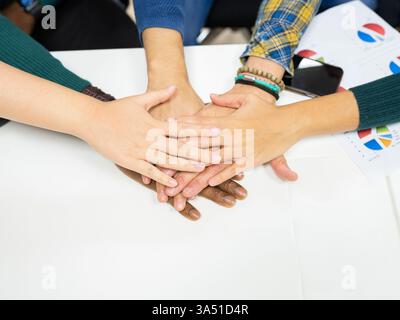 Vue de dessus d'un groupe diversifié de collègues autour d'une table, avec des graphiques et des mains empilés ensemble dans un geste de travail d'équipe. La scène capture la collaboration et la culture d'entreprise ou de start-up dans un environnement inclusif. Idéal pour les affaires, le leadership et les matériaux de consolidation d'équipe. Banque D'Images