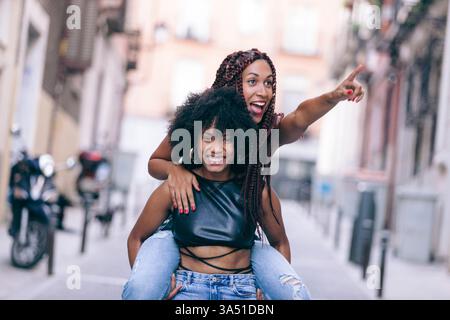 Joyeuse femme noire pointe et regarde loin tout en chevauchant son ami hispanique dans une rue ensoleillée. Un moment en plein air dynamique qui capte l'amitié et la joie interculturelle. Idéal pour les campagnes lifestyle et multiculturelles avec espace pour la messagerie. Banque D'Images