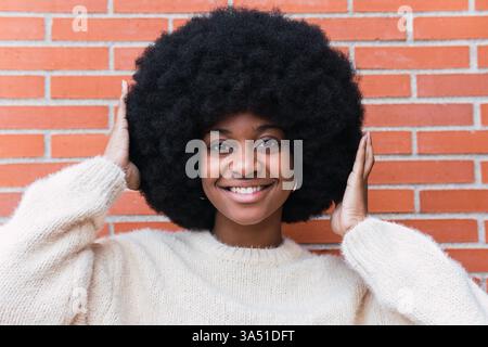 Femme noire souriante avec un afro se tient contre un mur de briques, les mains sur ses cheveux dans un portrait avec fond urbain. Cette image de style de vie dynamique convient à la beauté, à la mode et au contenu de style urbain confiant. Banque D'Images