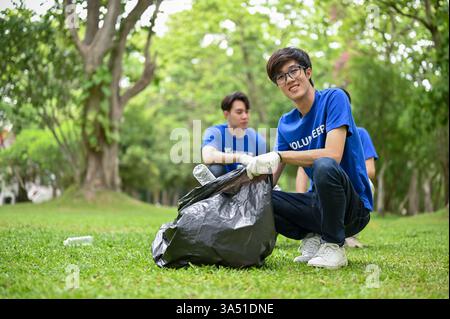 Un jeune volontaire asiatique heureux avec un sac poubelle en plastique nettoie un parc public avec son équipe. L'image met en valeur le service communautaire et la protection de l'environnement. Idéal pour le bénévolat, la RSE et les campagnes de sensibilisation à l'environnement. Banque D'Images