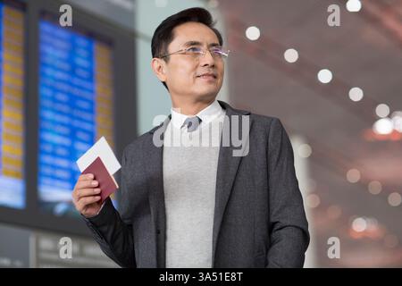 Souriant homme d'affaires chinois d'âge moyen regardant loin tenant passeport et billet debout à l'aéroport Banque D'Images
