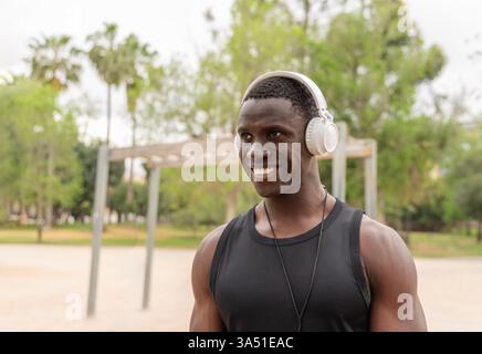 Homme noir souriant en vêtements de sport debout dans un parc pendant la journée, portant des écouteurs. La pose énergique et le cadre extérieur frais conviennent aux campagnes de fitness, de bien-être et de style de vie. Parfait pour la musique, le sport et l'image de marque orientée jeunesse. Banque D'Images