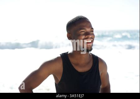 Gros plan portrait d'un homme afro-américain heureux souriant à la plage. Il est en plein air dans un cadre ensoleillé en bord de mer, rayonnant de confiance et de détente. Cette image optimiste fonctionne bien pour les campagnes de style de vie, de voyage et de bien-être. Banque D'Images