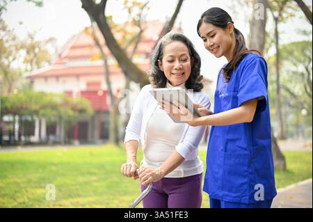 Souriante aidante asiatique dans un uniforme bleu montre une tablette à un patient âgé dans un parc, en utilisant une canne à quad à proximité. Le moment de la santé en plein air met en lumière les soins aux personnes âgées, le soutien infirmier et la communication innovante avec les patients. Utile pour les soins, les services de santé et les fonctions de bien-être des seniors. Banque D'Images