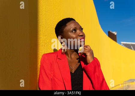 Femme noire souriante en costume rouge et boucles d'oreilles dorées touchant le menton et regardant loin tout en se tenant debout et appuyée sur le mur jaune sur la rue le jour ensoleillé Banque D'Images