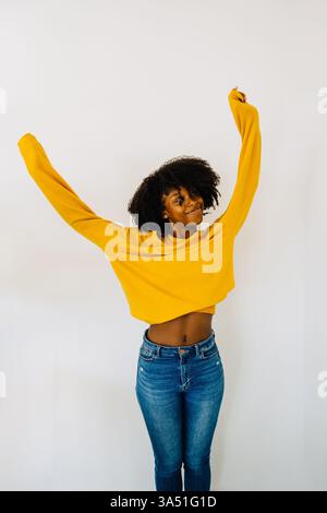 Femme hispanique souriante en denim et un pull jaune se tient à l'intérieur près d'un mur blanc avec les bras tendus. Ses cheveux bouclés et son style décontracté donnent un portrait gai de son style de vie. Banque D'Images
