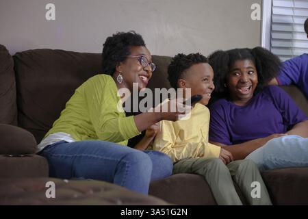 Mère Noire joyeuse avec un Afro est assise sur le canapé avec ses enfants, regardant la télévision et profitant d'un moment en famille. Cette scène intérieure chaleureuse est idéale pour les parents, la vie de famille et les campagnes de divertissement à domicile. Banque D'Images