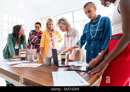 Un groupe diversifié de femmes d'affaires se réunissent autour d'une table avec un ordinateur portable et des documents, collaborant sur des plans d'affaires dans un bureau contemporain. La scène met en évidence le travail d'équipe, la diversité et l'autonomisation professionnelle pour les entreprises, les start-ups ou les femmes dans les thèmes d'affaires. Une image soignée et moderne adaptée aux contextes d'affaires et de réseautage. Banque D'Images