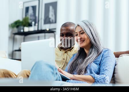 Un couple interracial se détend à la maison, souriant en regardant quelque chose sur un ordinateur portable. La scène du salon capture des moments chaleureux de style de vie d'amour, de connexion et de technologie partagée. Banque D'Images