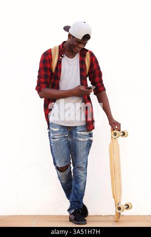 Portrait complet du corps d'un jeune homme noir heureux avec une planche à roulettes regardant son téléphone portable et souriant. Le style décontracté et moderne transmet la jeunesse, la mode et un style de vie actif. Convient pour le style de rue, le style de vie adolescent et la photographie urbaine. Banque D'Images