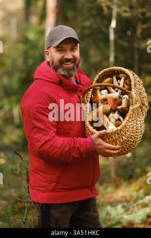 Un homme portant une veste rouge se tient dans une forêt vibrante, montrant fièrement un grand panier en osier rempli d'un assortiment de champignons récoltés Banque D'Images