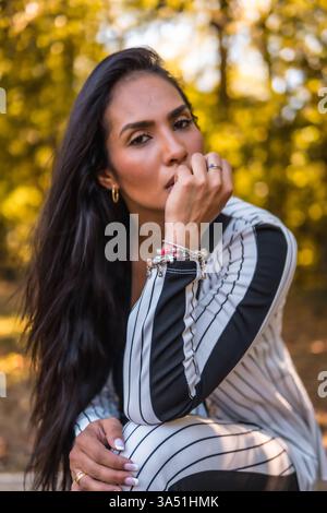 Portrait d'une jeune femme Latina élégante assise dans un parc, portant un élégant costume blanc avec des rayures noires. Le cadre estival et l'expression confiante traduisent un style de vie chic et une sophistication décontractée. Banque D'Images