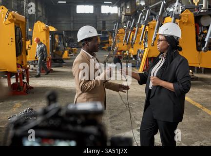 Une journaliste noire portant un casque de sécurité interviewe un ingénieur noir dans un entrepôt d'usine pendant la journée. La scène met en lumière les reportages sur site pour l'actualité de la fabrication et de l'industrie avec un microphone à main. Idéal pour les fonctionnalités sur les équipes d'ingénierie, les installations de production et les diffusions sur le terrain. Banque D'Images