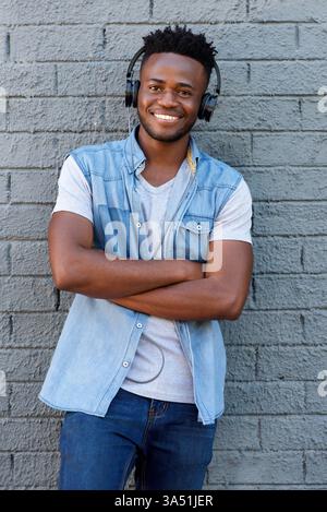 Smiling Black Man avec un court afro et barbe écoute de la musique sur des écouteurs contre un mur de briques grises. Le portrait urbain décontracté rayonne de confiance et de loisirs pour les campagnes de style de vie et de musique. Idéal pour le style urbain et les images contemporaines. Banque D'Images