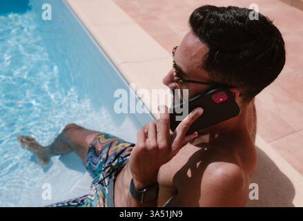 Homme hispanique assis au bord de la piscine parlant sur le téléphone portable en été Banque D'Images