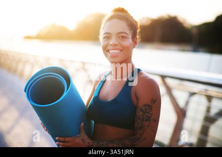 En route vers l'entraînement. Jeune femme tatouée souriant tout en tenant un tapis de sport et en se dirigeant vers l'entraînement Banque D'Images
