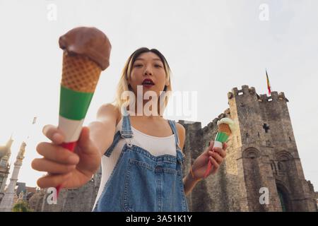 Femme asiatique appréciant un cône de crème glacée sucrée dans une rue urbaine ensoleillée. Elle savoure un cornet de gaufres, une friandise estivale rafraîchissante idéale pour les campagnes alimentaires et lifestyle. Ce moment extérieur franc ajoute un accent savoureux et coloré aux scènes de la ville. Banque D'Images