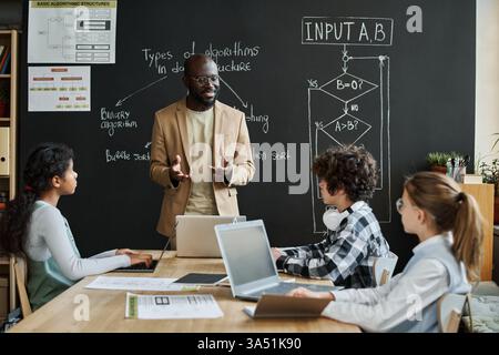Un enseignant afro-américain explique les concepts d'interface informatique aux enfants lors d'une leçon DE TI. Les élèves s'assoient à une table avec des ordinateurs portables dans une salle de classe. Idéal pour les technologies de l'éducation, l'alphabétisation numérique et l'imagerie d'apprentissage en classe. Banque D'Images