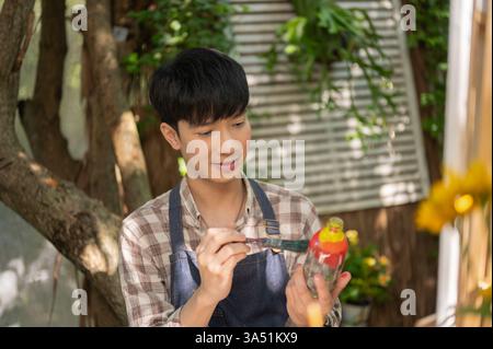 Un jeune homme asiatique positif dans un tablier rejoignant un atelier d'art, peignant une bouteille en verre, fabriquant à la main une bouteille décorative. Concept de passe-temps Banque D'Images