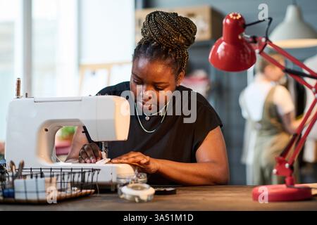 Jeune tailleur noire utilisant une machine à coudre électrique dans un atelier occupé. Le cadre intérieur montre un collègue à proximité, mettant en évidence la couture professionnelle, la couture et la production de mode. Idéal pour le contenu sur les femmes dans l'artisanat, la mode petite entreprise, et couturières qualifiées. Banque D'Images