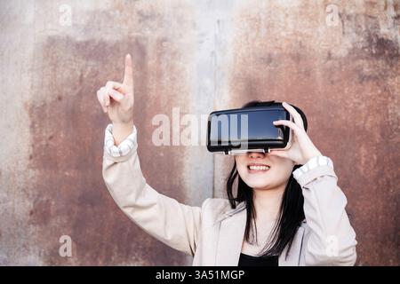 Femme asiatique souriante portant un casque VR pointe vers le haut, debout contre un mur minable dans la journée. Cette image met en évidence la technologie moderne, la réalité virtuelle et les concepts de métaverse, ce qui la rend adaptée aux contextes de technologie, d'innovation et de mode de vie numérique mettant en vedette un sujet asiatique. Banque D'Images
