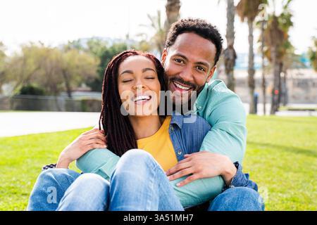 Couple hispanique souriant embrassant sur l'herbe dans un parc de Barcelone pendant la journée. Un moment joyeux capturé en extérieur dans un cadre ensoleillé. Idéal pour le style de vie, la romance et les thèmes d'activités de plein air. Banque D'Images