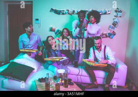 Scène vibrante d'un groupe diversifié de jeunes amis célébrant une fête à la maison. Dans un appartement confortable aux lumières chaudes, ils partagent des toasts, des collations et des rires, capturant un moment de vie insouciant. Banque D'Images