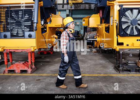 Portrait d'une jeune technicienne en salopette, casque de sécurité et gants, se déplaçant parmi de grandes machines industrielles dans un entrepôt très fréquenté. La scène met en évidence les métiers qualifiés et les environnements de fabrication. Adapté aux images de sécurité de l'ingénierie, de l'usine et du lieu de travail. Banque D'Images