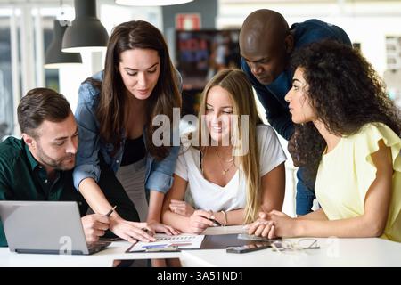 Groupe diversifié de professionnels en tenue décontractée assis et penché en avant à une table, collaborant dans un bureau. Cette image est parfaite pour le travail d'équipe, le brainstorming et les réunions d'affaires dans des environnements de travail modernes. Banque D'Images