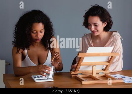 Femme hispanique mélangeant l'aquarelle à côté d'un ami caucasien à une table avec un chevalet contre un mur gris. Parfait pour les loisirs créatifs, l'amitié et les activités artistiques d'intérieur. Banque D'Images